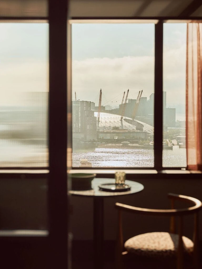 Sunlight pours through the guest-room windows towards the Thames and Greenwich Peninsula, framing the O2’s distinctive roofline and cranes beyond. The view is filtered by dark walnut framing and reeded glass, turning London’s waterside skyline into part of Holloway Li’s carefully composed interior.