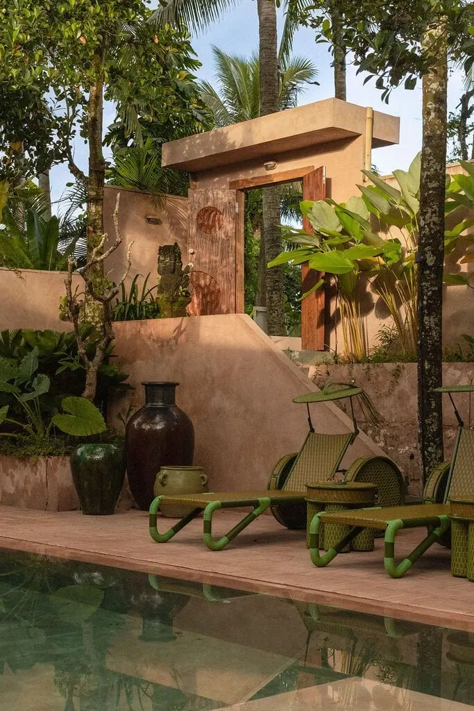 Poolside at Hotel Moon Gang, lime-green woven sun loungers with parasol-like visors sit on a terracotta deck beside glazed green and dark ceramic urns. Behind, a rendered gateway with a carved timber door punctured by a circular motif leads up a ramp, framed by palms and a weathered stone Balinese statue.