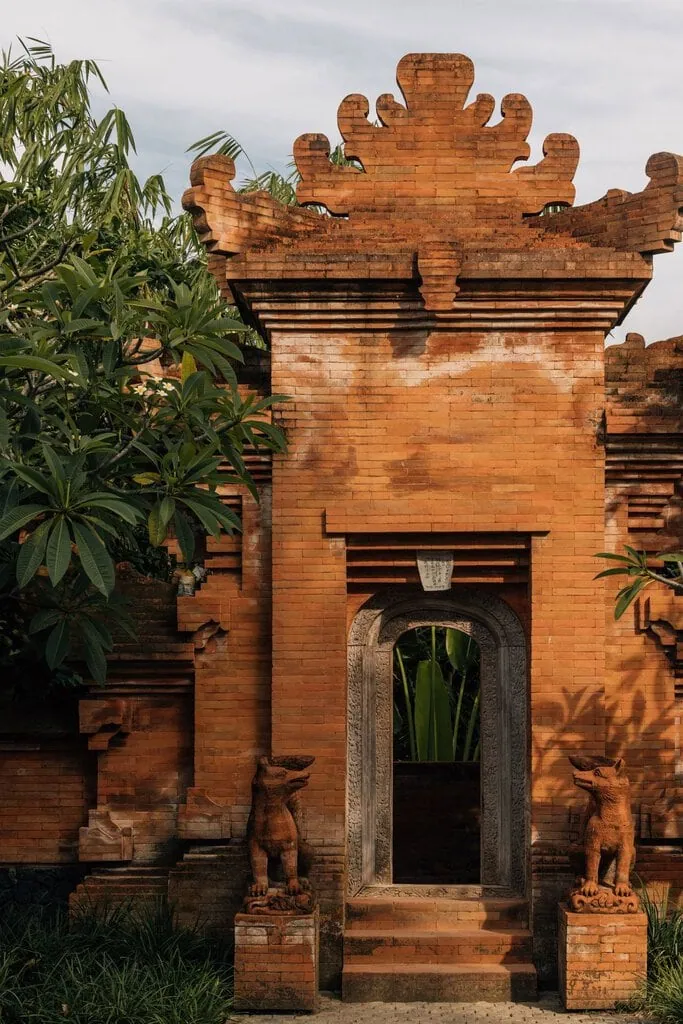Warm evening light gilds the brick garden temple, its stepped terracotta gateway crowned by an elaborate sculpted crest and flanked by carved guardian creatures on plinths. A narrow arched doorway frames tropical greenery, frangipani leaning in at left, the weathered masonry glowing against a soft grey sky.