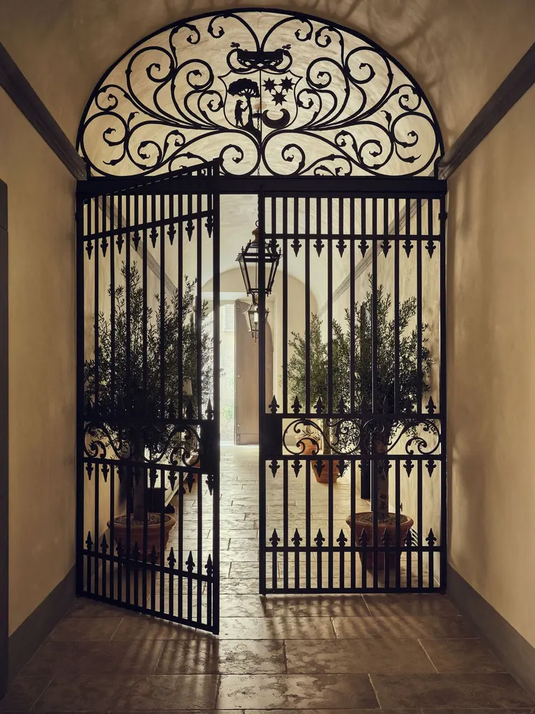 A wrought-iron gate at La Réserve Firenze, crowned by a scrolling lunette with a heraldic shield of a tree, stars and a crescent, stands half open onto the entrance passage. Beyond the bars, olive trees in terracotta pots and hanging lanterns lead the eye towards daylight from the street.