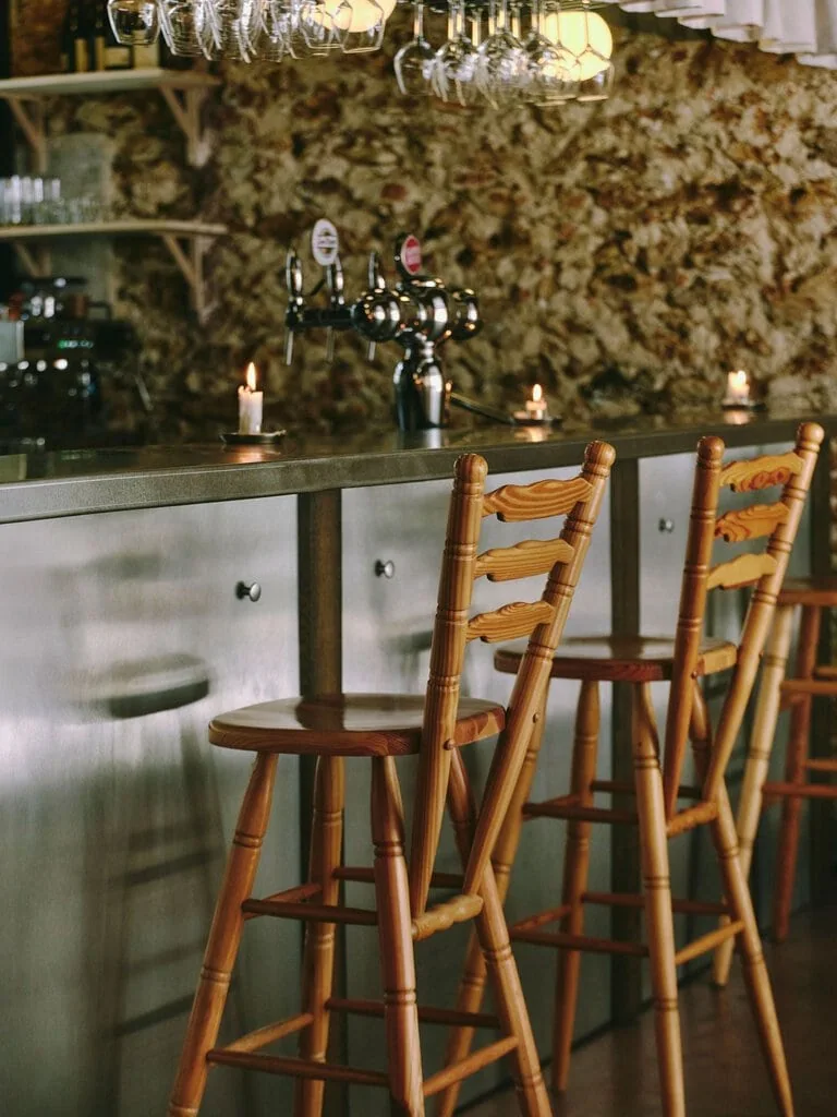 Pine ladder-back stools with carved rails and turned legs pull up to the brushed-steel bar at SaltoBar in Paris. Behind them, chrome beer taps and stubby candles stand against a rough rubblestone wall. Rows of inverted wine glasses hang from racks overhead, catching the glow of spherical lamps.