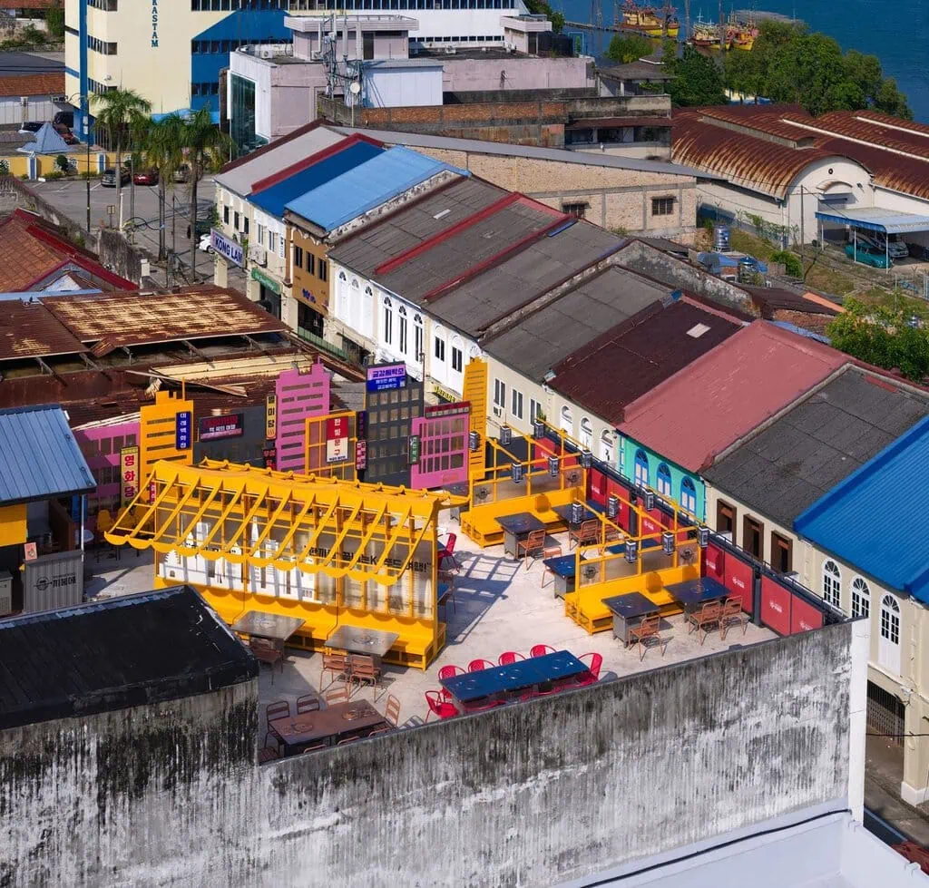 Seen from above, JUJURWORK's rooftop scheme reveals its full composition: the crisp orange hanok canopy, a stylised skyline screen of pink, black and lettered lightboxes, and colour-blocked stainless-steel barbecue tables. Kuantan's old shophouse roofs and Jalan Besar's multicultural fabric press in on every side.