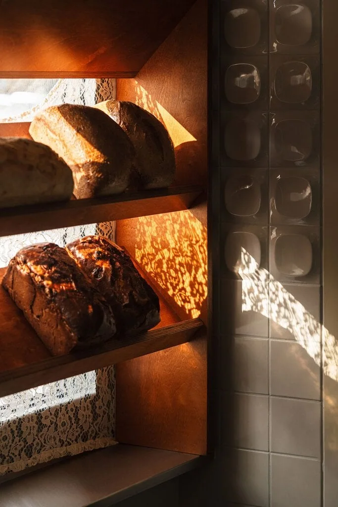 Sunlight filters through lace curtains onto loaves ranged across plywood shelves at Węzeł Cieplny, casting lace shadows over the timber and across the glossy white 3D tiles alongside. The warm grain and dimpled ceramic surfaces meet in a study of texture and pattern.