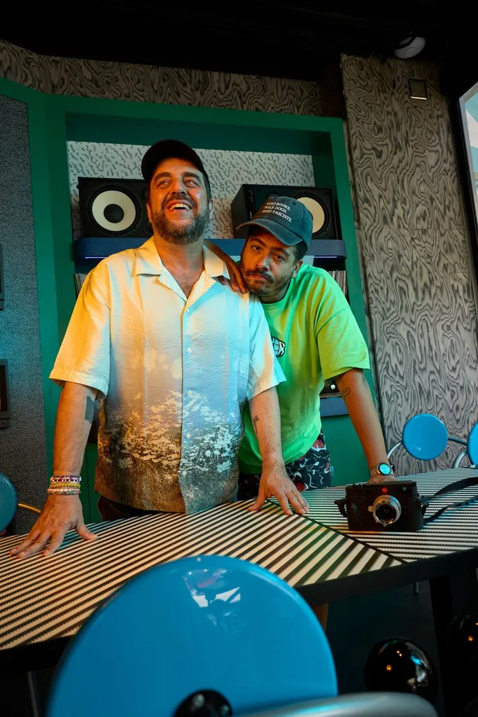 The two founders of Seth & Manni's pose at a striped bistro table, one resting his chin on the other's shoulder. Behind them, a teal-lacquered alcove holds speakers and stacked records against Memphis-patterned backing, framed by grey wood-grain veneer. In the foreground, the glossy blue disc of a First chair looms large.