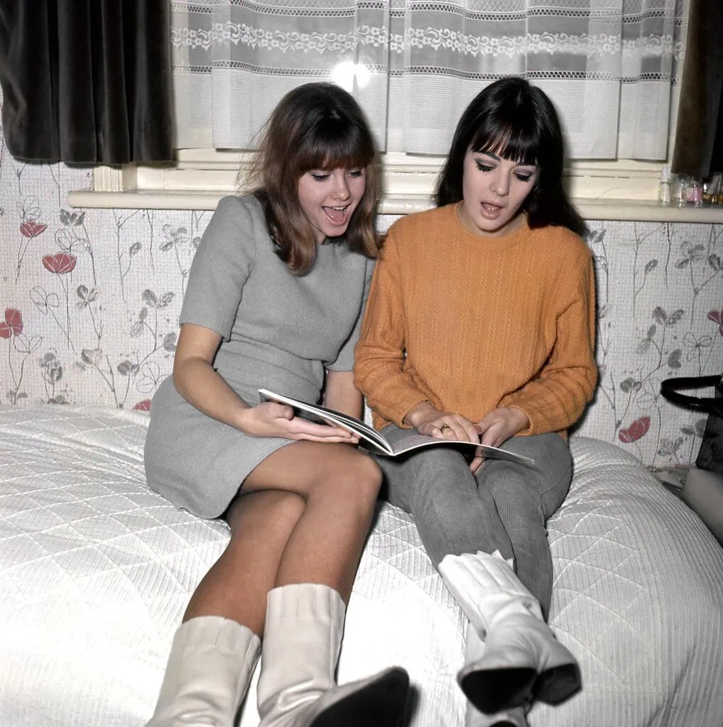 Olivia NewtonJohn left with Pat seated on bed floral wallpaper window with lace curtains