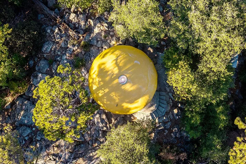 an aerial view shows the yellow Futuro house nestled among trees and rocky terrain in Idyllwild, California