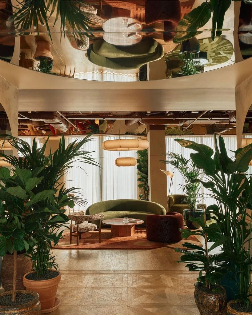 Lush planting frames the entrance lounge at Locke London Canary Wharf, where Holloway Li layers curved mid-century seating, patterned rugs and warm timber beneath a mirrored ceiling. The reflective surface doubles the greenery and furnishings overhead, lending the relaxed, biophilic setting an unexpected sense of theatricality.
