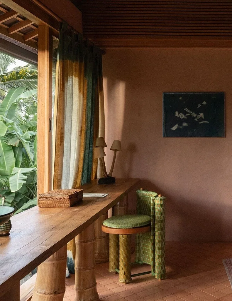 A suite study corner, where a raw-timber desk on bamboo-style legs meets a green woven tub chair beneath a dark abstract artwork. Plant-dyed curtains in green and ochre filter jungle light, a cluster of shaded bamboo lamps and a woven box lending the pink-plastered nook a quiet, crafted calm.