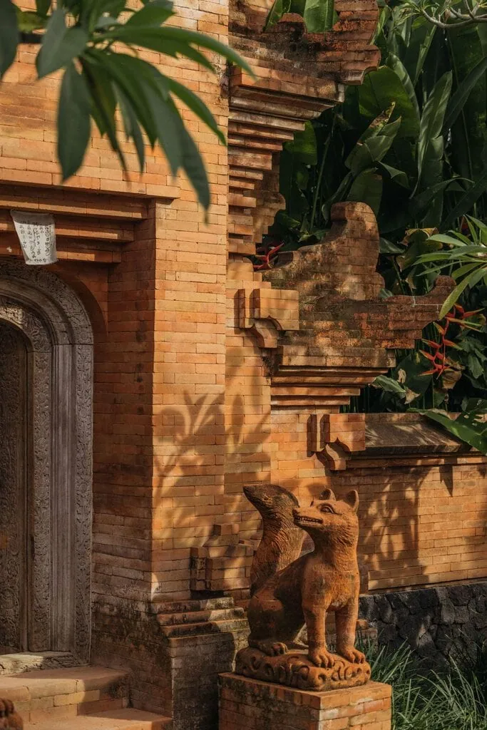 A detail of the brick entrance gate catches golden light on its finely laid terracotta courses and stepped, sculpted forms. A pair of carved guardian creatures sits sentinel beside a richly ornamented timber door, tropical foliage and red heliconia framing the warm, weathered masonry.