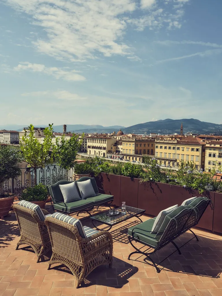 Under a wide summer sky, the rooftop terrace of La Réserve Firenze gathers wrought-iron and wicker seating on a herringbone terracotta floor. Beyond the planted parapet and an iron balustrade, the Arno and its lungarno palazzi lead the eye towards the Tuscan hills.