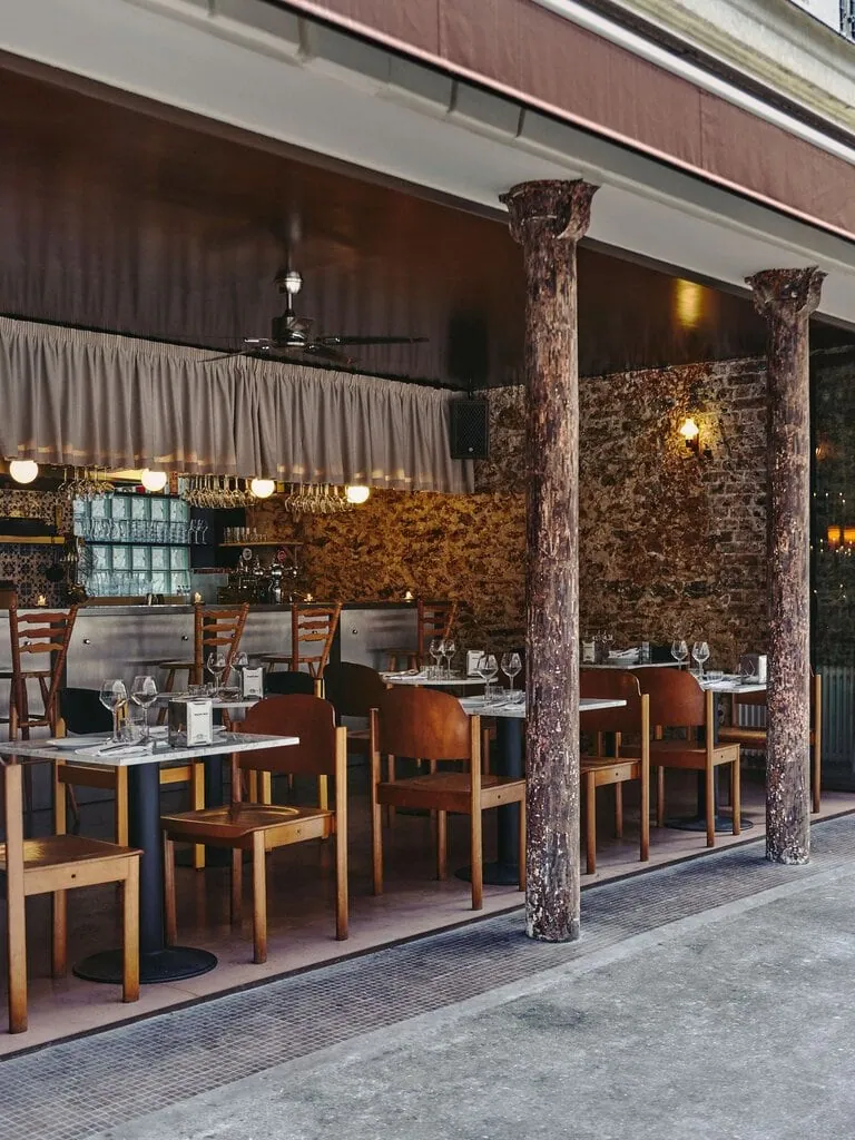With its glazed frontage fully open, SaltoBar in Paris spills towards the pavement between two weathered cast-iron columns. Marble tables and warm wooden chairs sit on a mosaic-tiled threshold. Beyond them, the steel bar, pleated valance and glowing globe lights stand against a textured rubblestone wall.