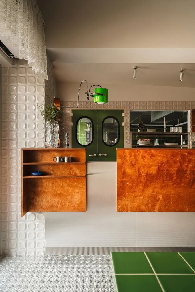 The bar at Węzeł Cieplny, where stained plywood panels meet glossy white 3D tiles with dimpled square reliefs. Green porthole doors lead to the kitchen, a green enamel task lamp arcs overhead on a chrome stem, and the green tiled communal table enters at the lower right.