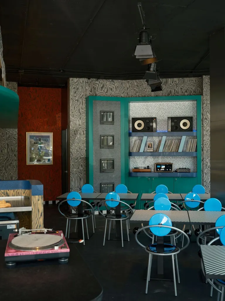 A pink turntable in the foreground sets the tone for the dining room at Seth & Manni's, where Michele De Lucchi’s blue disc-backed First chairs gather around striped tables. Beyond, the teal media wall of monitors, speakers and records meets grey and rust-red wood-grain walls. Theatrical spotlights hang from the black ceiling.