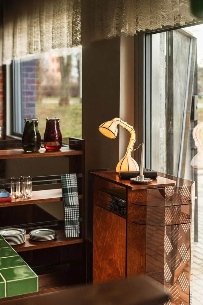 Beside the glazed entrance at Węzeł Cieplny, a stained plywood host stand with a smoked polycarbonate side panel carries a sculptural table lamp and a candle. Lace curtains frame the window, while preserving jars, tumblers and stacked plates fill the plywood shelving alongside the green tiled table.