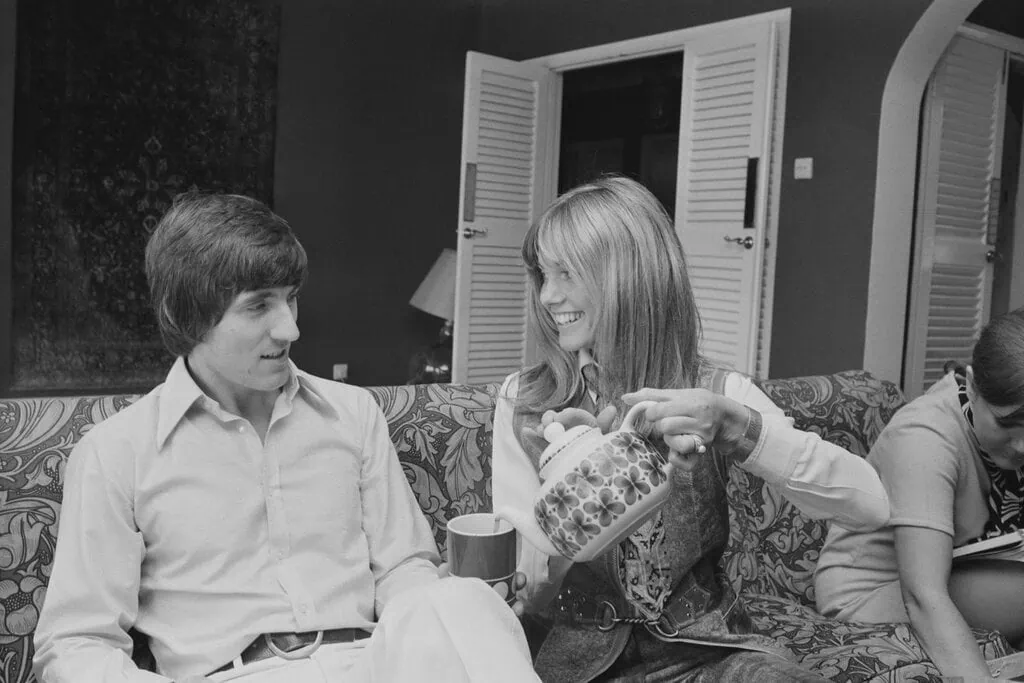blackandwhite photo of Olivia NewtonJohn right and Bruce Welch at home on sofa pouring tea