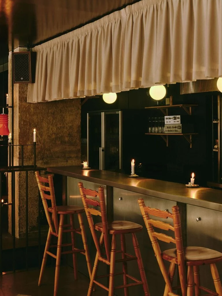 The bar at SaltoBar in Paris after dark, where turned-pine ladder-back stools pull up to a brushed stainless-steel counter dotted with candles. Above, a ruffled fabric valance hangs over opal globe lights. Vintage hi-fi components rest on wall shelves, and a wood-cased speaker is mounted on a textured concrete pillar.