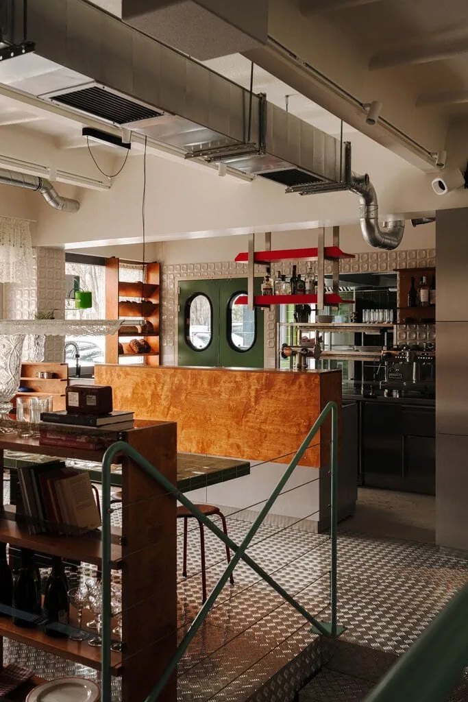 Looking across Węzeł Cieplny towards the bar, where a stained plywood counter meets glossy white 3D tiles and green porthole doors. Exposed ductwork runs overhead, mint-green railings cross the foreground, and lace fabric drapes over a tubular pendant above the communal table.