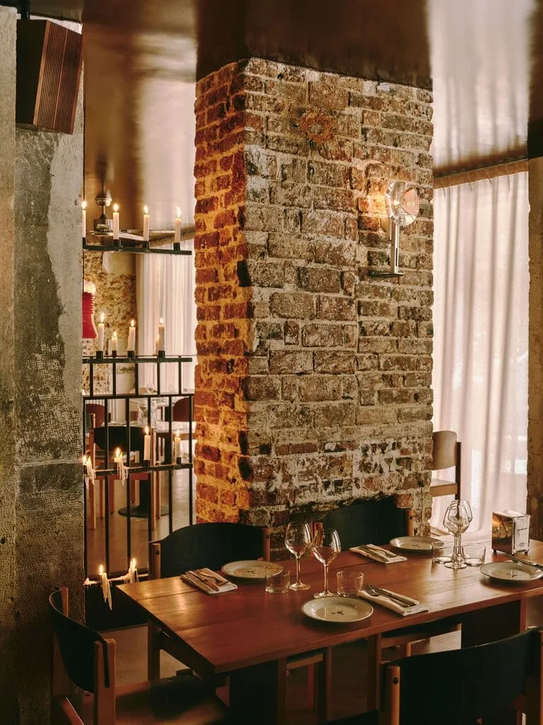 A timber table set for dinner nestles against the base of a rugged brick pillar at SaltoBar in Paris. A glass sconce catches the light on the weathered masonry. To the left, tiers of dripping candles on a black metal screen glow against raw concrete. Sheer curtains diffuse the daylight behind.