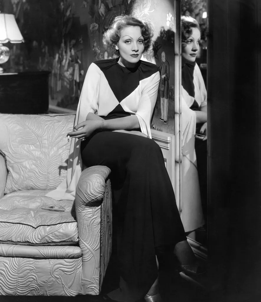 blackandwhite photo of Marlene Dietrich sitting on arm of chair at home mirror to right