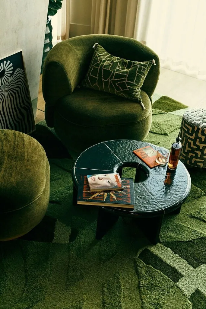 A close-up of the ground-floor lounge highlights its tactile layering of moss-green upholstery, sculptural furniture and a deeply textured geometric rug. A glossy black coffee table, patterned textiles and graphic artwork sharpen the retro palette, while filtered daylight gives this intimate seating zone a relaxed, residential character.