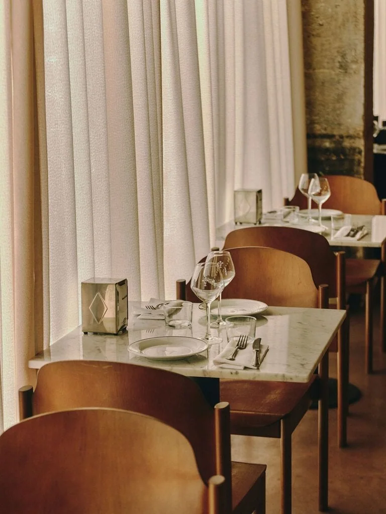 Sheer curtains filter the daylight along the window tables at SaltoBar in Paris, casting a soft, peachy glow over white marble tops. Curved plywood chairs in warm wood tones sit beside steel napkin dispensers with an engraved diamond motif. Wine glasses etched with the SaltoBar name complete the table settings.
