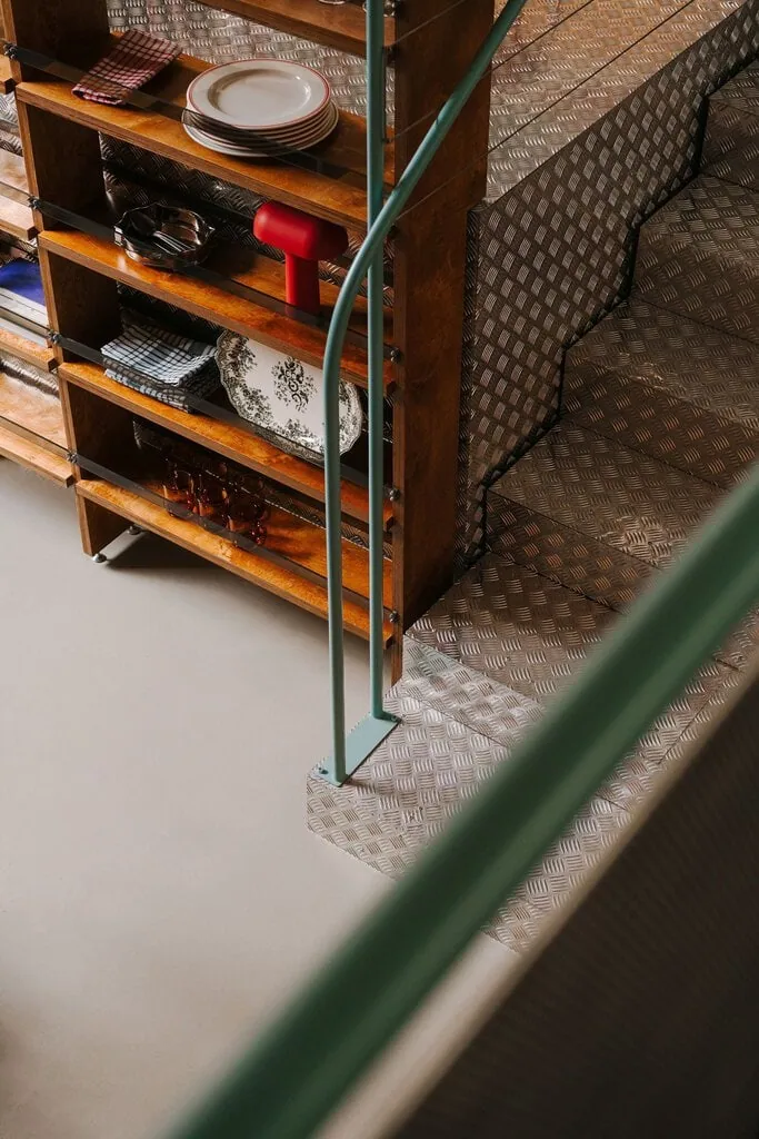 A detail at Węzeł Cieplny where mint-green railing tubes rise from a tread plate base beside plywood shelving stocked with rimmed plates, checked linen, a floral platter and amber glasses. A red portable table lamp punctuates the timber, while tread plate wraps the adjacent steps.