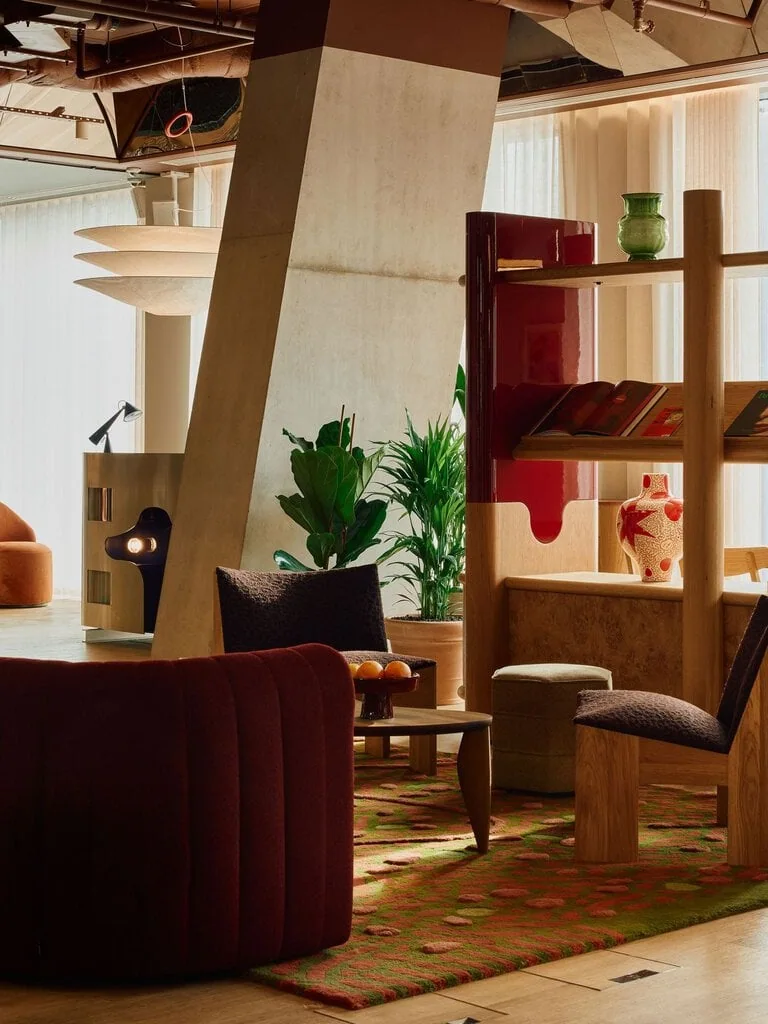 A richly layered lounge vignette brings together burgundy upholstery, patterned rugs, sculptural timber furniture and Holloway Li’s glossy red-lacquer joinery. The angular concrete column and exposed ceiling temper the retro warmth with a raw architectural edge, while the stainless-steel DJ booth glows in the background like a cinematic prop.