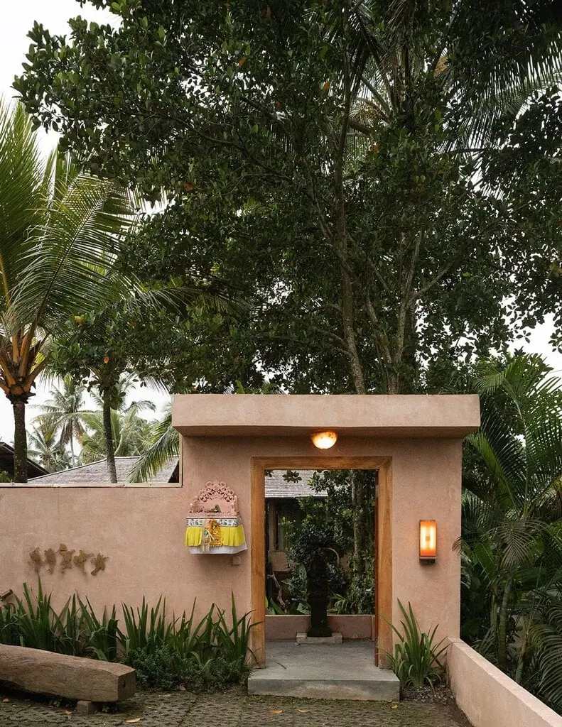 The entrance to Hotel Moon Gang, framed by a cinnamon-rendered gateway with a timber-lined doorway. A pink carved Balinese shrine draped in yellow cloth sits on the wall beside an amber sconce, while dense jungle foliage and coconut palms rise behind. A carved timber bench rests on the cobbled forecourt at dusk.