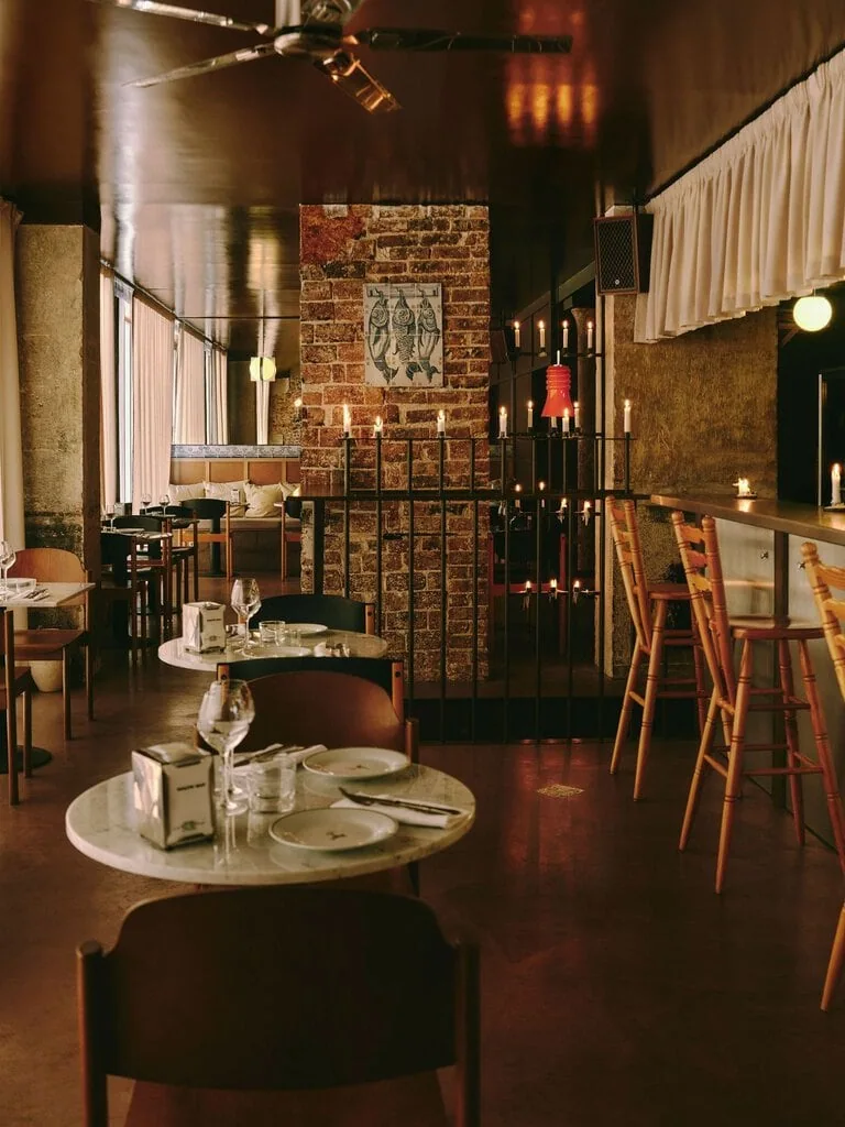 Round marble tables lead the eye through SaltoBar in Paris towards a brick pier hung with a tiled panel of three hanging fish. A black metal railing crowded with candles and a scarlet pendant separates the dining room from the bar. There, turned-pine stools line a steel counter beneath a pleated cream valance.