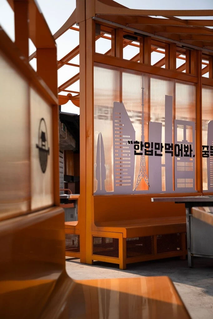A tight study of the central pavilion's corner, its upturned eaves and ribbed polycarbonate panels catching low sun. A cut-out skyline motif and Korean lettering sit across the translucent screen, while the pig-and-hanok logo appears on an adjacent panel, the whole bathed in warm amber light.