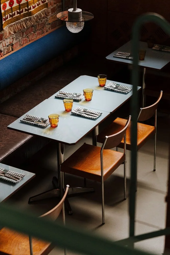 A pale blue laminate-topped table at Węzeł Cieplny set with checked napkins and amber tumblers, seen from above. Bentwood and chrome chairs sit alongside a brown leather and blue denim banquette, while a textured glass wall lamp on a cantilevered bracket casts light from the exposed brick wall.